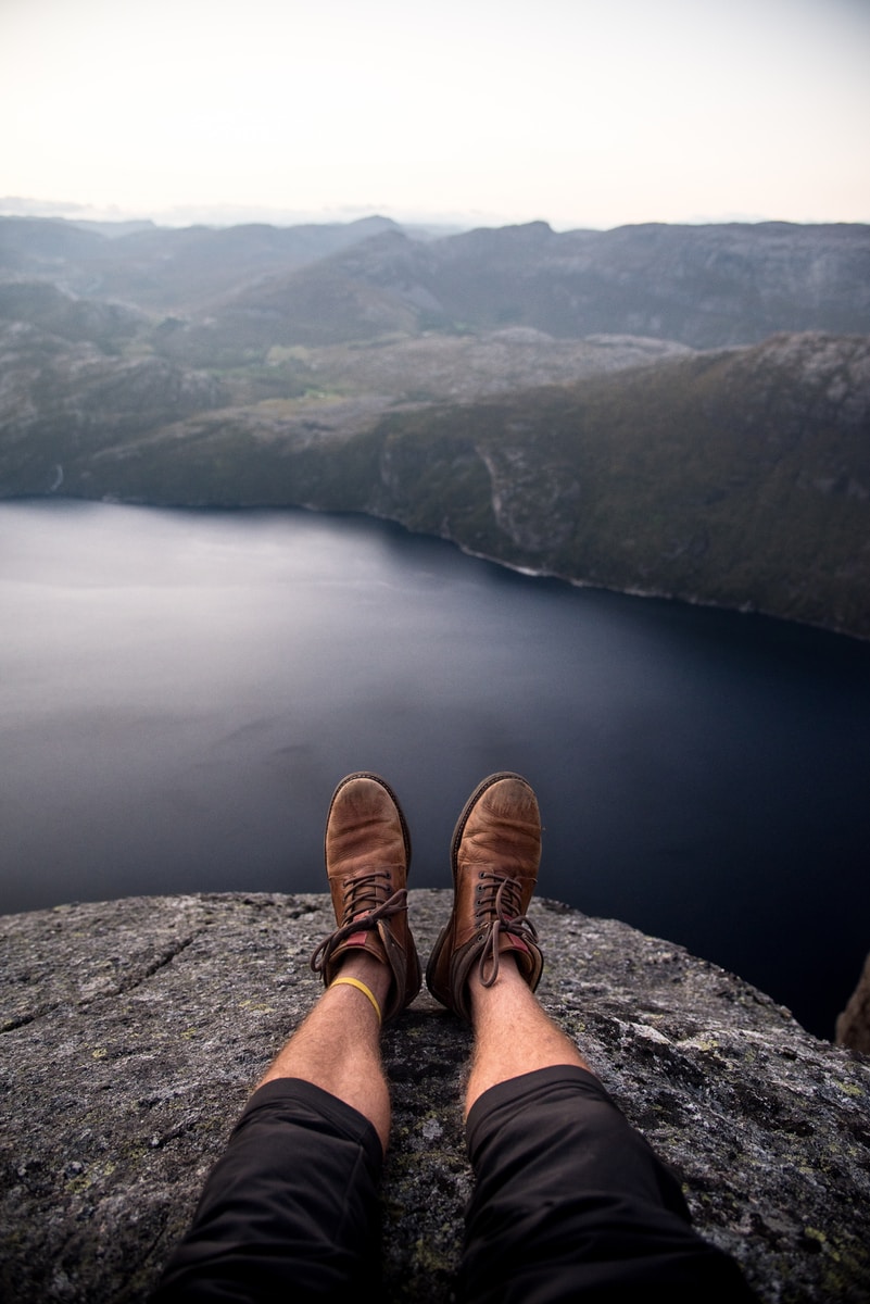person wearing brown boots sitting on cliff in front of grey calm body of water and rock formations during daytime