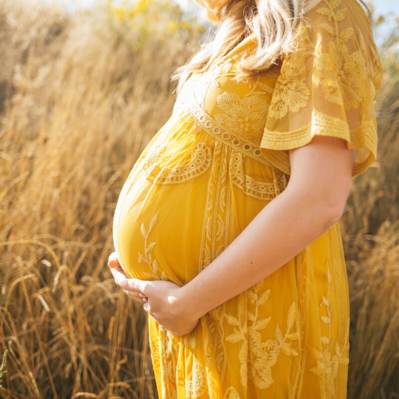 pregnant woman wearing yellow floral dress standing while touching her tummy and facing her right side near brown field during daytime